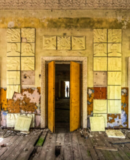 A wall in disrepair with three doors. The center door has a silhouette of a figure at the end of a hallway.