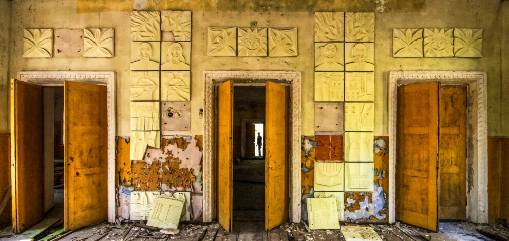 A wall in disrepair with three doors. The center door has a silhouette of a figure at the end of a hallway.
