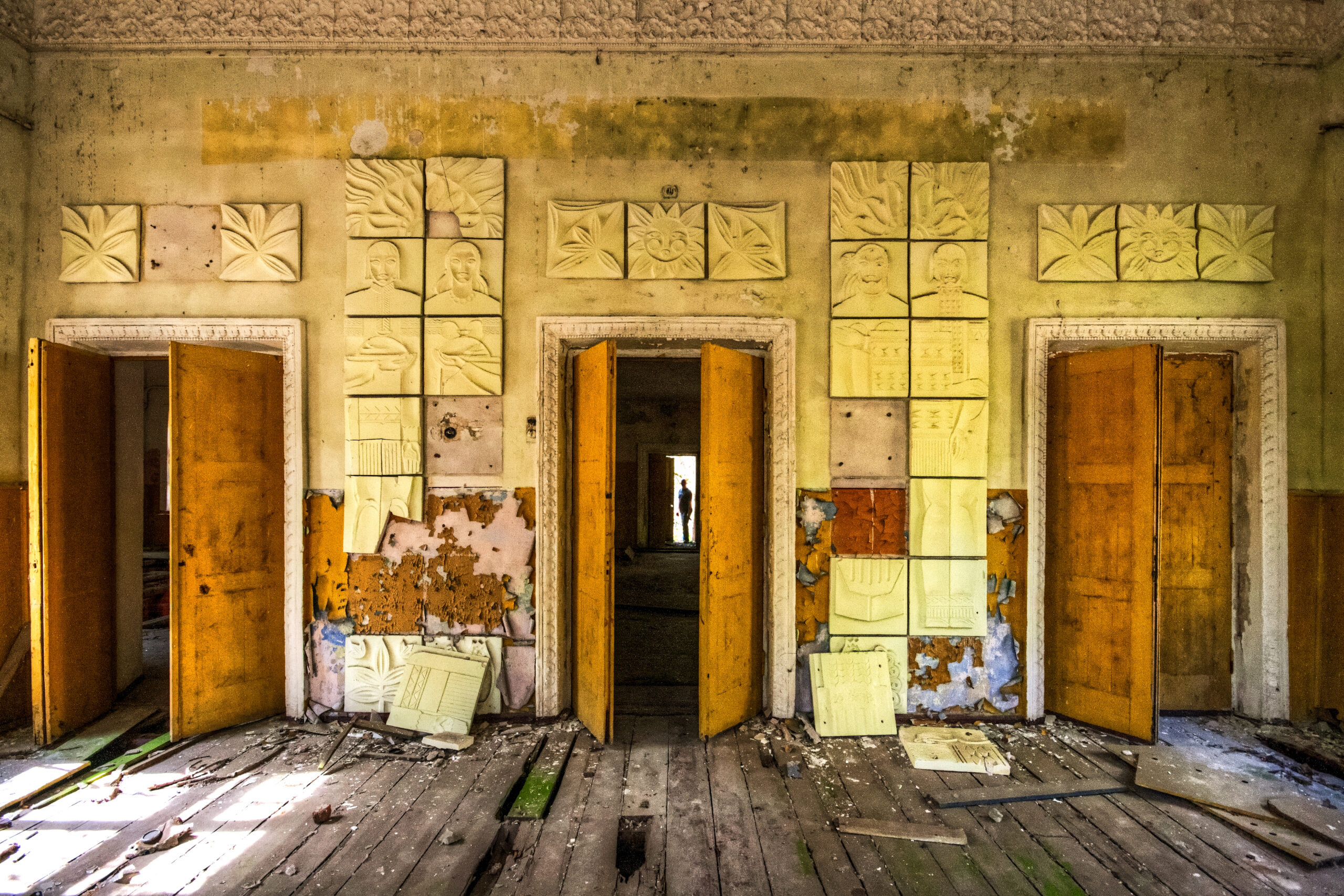 A wall in disrepair with three doors. The center door has a silhouette of a figure at the end of a hallway.