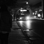 Bus at night in distance, stopped with passengers getting on, woman in shadow in foreground