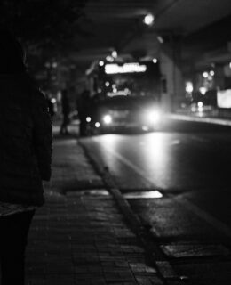 Bus at night in distance, stopped with passengers getting on, woman in shadow in foreground
