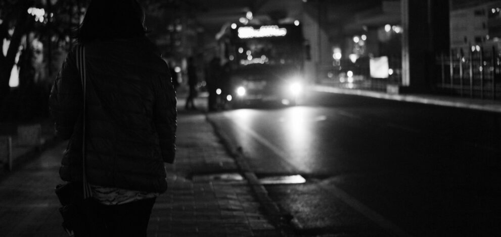 Bus at night in distance, stopped with passengers getting on, woman in shadow in foreground