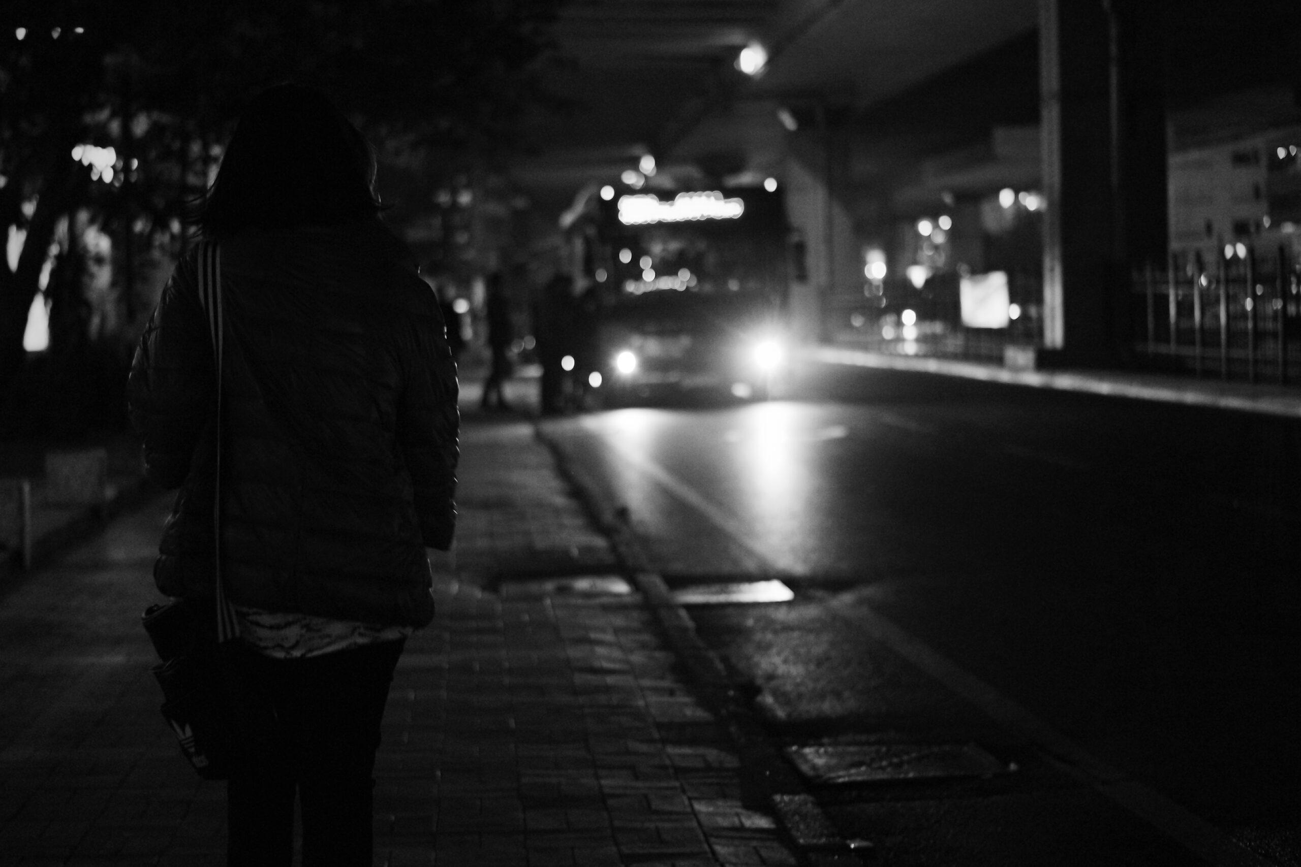 Bus at night in distance, stopped with passengers getting on, woman in shadow in foreground